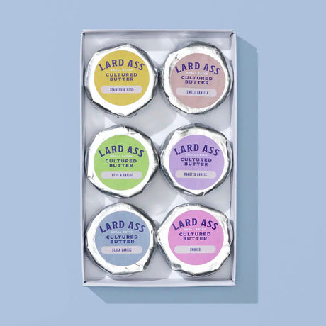 A boxed set of six Lard Ass Cultured Butter tins arranged in a 3-by-2 grid inside a clear plastic tray. The tins have silver foil lids with pastel-colored labels in yellow, pink, green, purple, blue, and lavender, set against a solid light blue background.