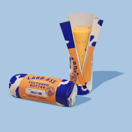 Two packaged wheels of Lardass Cultured Butter in the Chilli Lime flavor, shown on a light blue background. One wheel is lying flat, and the other is standing upright with its wrapper partially peeled back to reveal the orange-yellow butter inside.