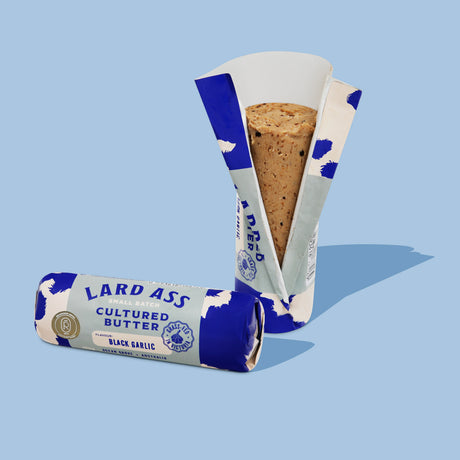 Two packages of Lard Ass Cultured Butter, one standing upright and one lying down, showing the blue and white cow-print packaging and a slice of the browned butter inside, set against a solid light blue background.