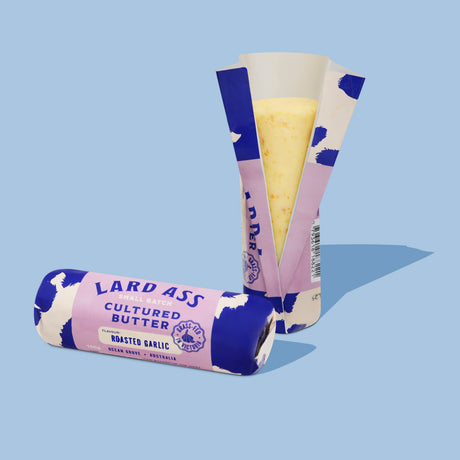 Two wrapped butter sticks on a light blue background. One is standing upright with its paper wrapper partially peeled back to show pale yellow butter inside, and the other lies on its side.