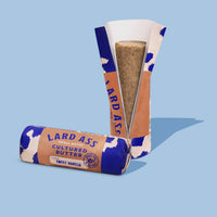 Two Lard Ass cultured butter sticks on a light blue background, one standing in its paper wrapper and one lying flat on its wrapper, with the standing stick cut open to show the vanilla-colored butter inside.