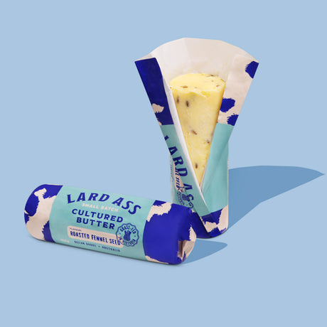 Two wrapped slices of Lardass Roasted Fennel Seed Cultured Butter on a light blue background, one standing upright and one lying horizontally.