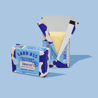 A bar of Lard Ass Unsalted Cultured Butter shown with its branded paper packaging on a light blue background. The packaging is blue and cream with a cow-print motif, and the butter itself is pale yellow and partially visible inside an open box.