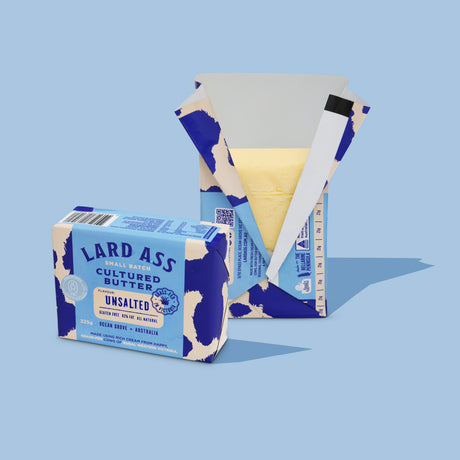 A bar of Lard Ass Unsalted Cultured Butter shown with its branded paper packaging on a light blue background. The packaging is blue and cream with a cow-print motif, and the butter itself is pale yellow and partially visible inside an open box.