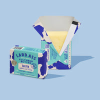 A package of Lard Ass Cultured Butter Salted shown with one closed carton in front and one open carton behind it revealing yellow butter inside, on a light blue background.