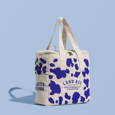 A canvas tote bag with a blue and white cow print pattern, featuring the brand name 'LARD ASS' printed on the front, set against a solid light blue background.