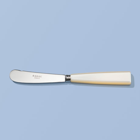 A SABRE Paris butter knife with a silver stainless steel blade and a cream-colored bone handle, displayed horizontally on a solid light blue background.