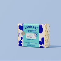 A single wrapped bar of Lard Ass Cultured Butter standing upright on a light blue background. The packaging is white with blue cow-print spots and a turquoise label reading 'LARD ASS CULTURED BUTTER' and 'CHILLI PEPPER'.