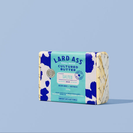 A single wrapped bar of Lard Ass Cultured Butter standing upright on a light blue background. The packaging is white with blue cow-print spots and a turquoise label reading 'LARD ASS CULTURED BUTTER' and 'CHILLI PEPPER'.