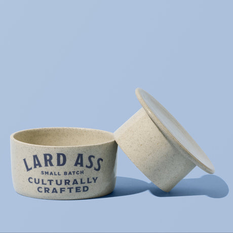 A small round ceramic container with its lid leaning against it on a light blue background. The container is off-white with dark blue text that reads 'LARD ASS SMALL BATCH CULTURALLY CRAFTED'.