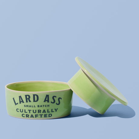 A light green ceramic lard tin with its lid leaning against it on a solid light blue background. The tin features the text 'LARD ASS SMALL BATCH CULTURALLY CRAFTED' in dark green lettering.