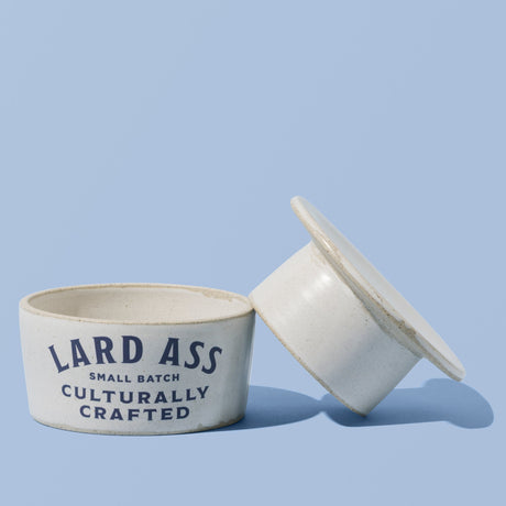 A small white ceramic container with its lid leaning against it on a light blue background. The container is printed with the text 'LARD ASS SMALL BATCH CULTURALLY CRAFTED' in dark blue lettering.