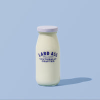A glass bottle filled with white milk or yogurt, featuring a white lid and blue text that reads 'LARD ASS CULTURALLY CRAFTED', set against a solid light blue background with a soft shadow.