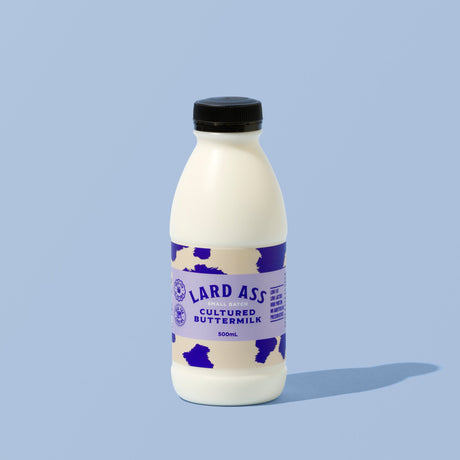 A 300ml bottle of Lard Ass Cultured Buttermilk with a white body, purple cow-print label, and black cap, set against a solid light blue background.