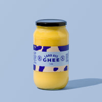 A glass jar of Lard Ass Ghee with a black lid and a white label featuring blue cow-print patterns, set against a solid light blue background.