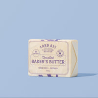A single stick of Lard Ass Reshaped Baker's Butter stands upright on a light blue background. The butter stick is wrapped in cream-colored paper with purple text and decorative borders, and a small gold foil seal is visible on the right side.